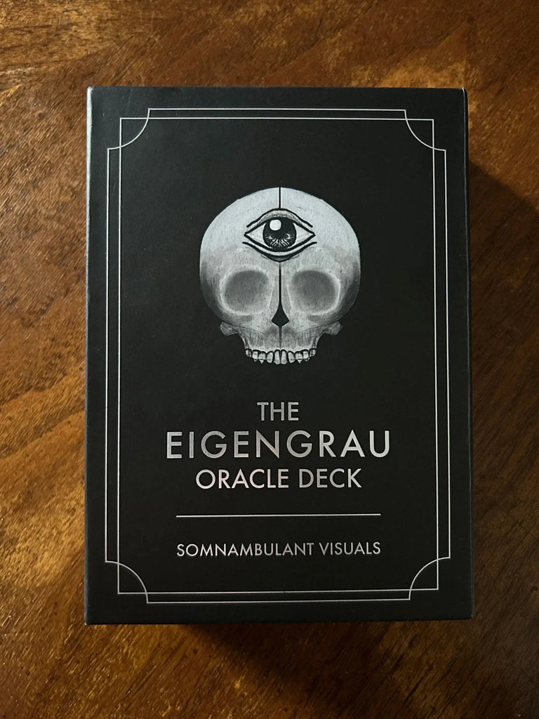 The Eigengrau Oracle Deck box displayed flat on a warm wooden surface, featuring a black cover with a grayscale skull illustration and a third eye symbol, with silver text and decorative corner border
