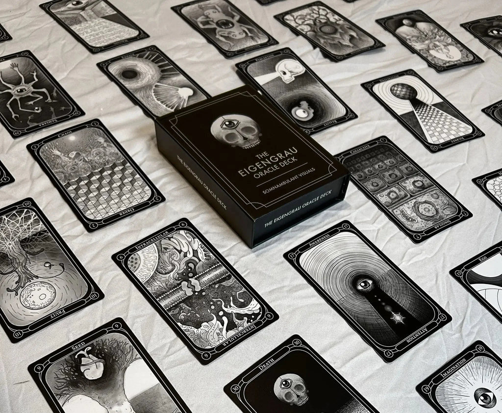 Black and white oracle deck box labeled 'The Eigengrau Oracle Deck' centered among fanned tarot cards with intricate skull and geometric illustrations on a cream fabric background.