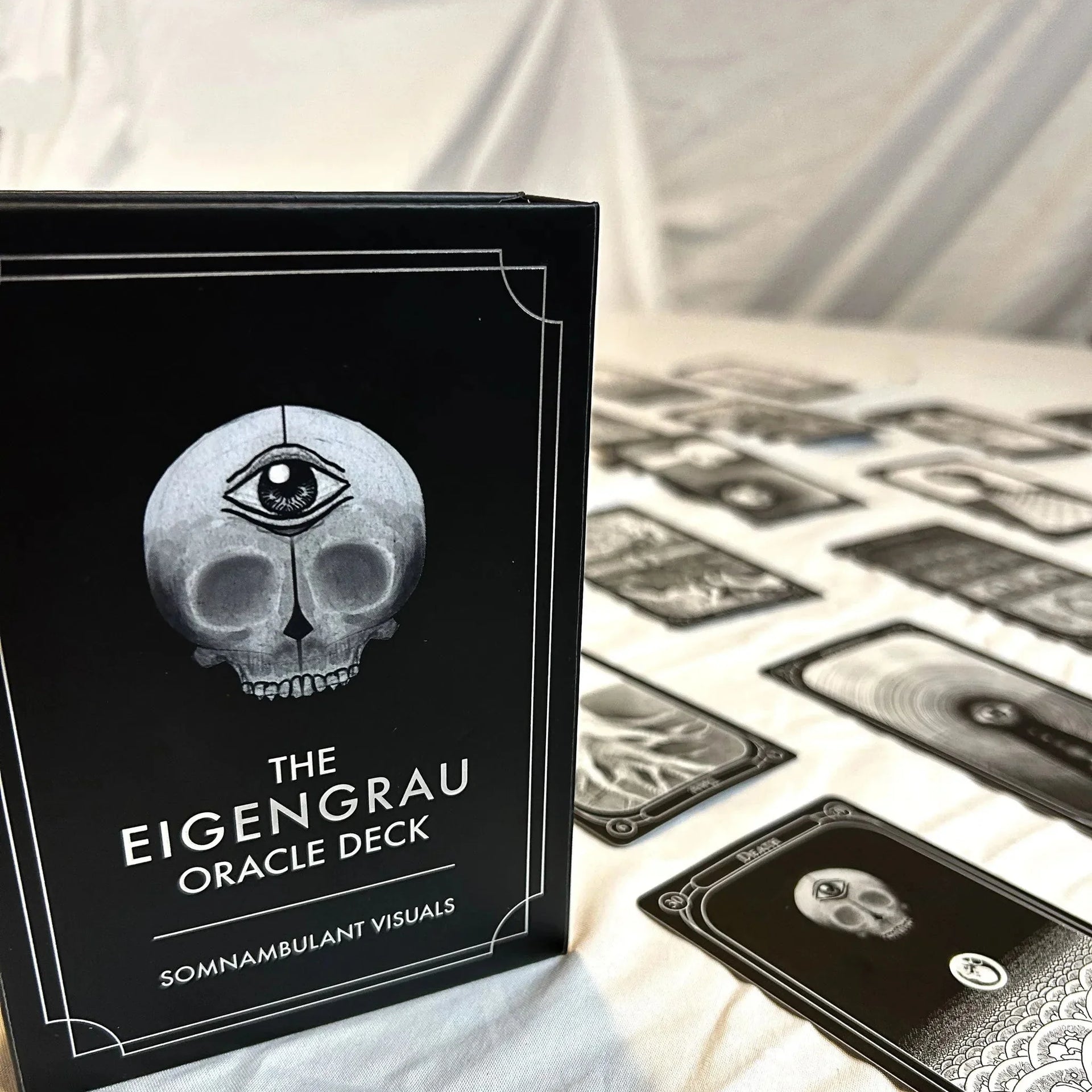 The Eigengrau Oracle Deck box featuring a skull with a third eye sits beside fanned tarot cards on crumpled white fabric, showcasing the dark monochromatic aesthetic of the deck.