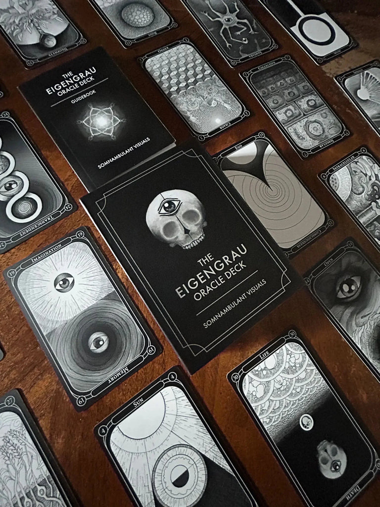 The Eigengrau Oracle Deck guidebook and black-and-white oracle cards arranged on a rich wooden surface, displaying intricate mystical imagery including skulls, eyes, and geometric patterns.