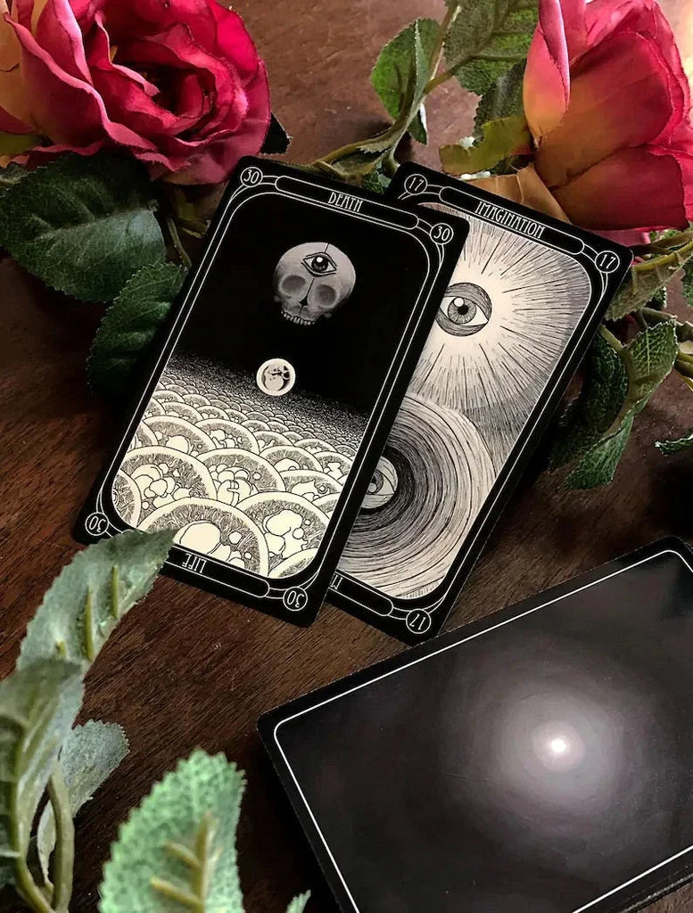 Two black tarot cards with intricate white line art depicting a skull and a spiraling eye design, arranged on a dark wood surface surrounded by deep pink roses and green foliage beside a dark crystal