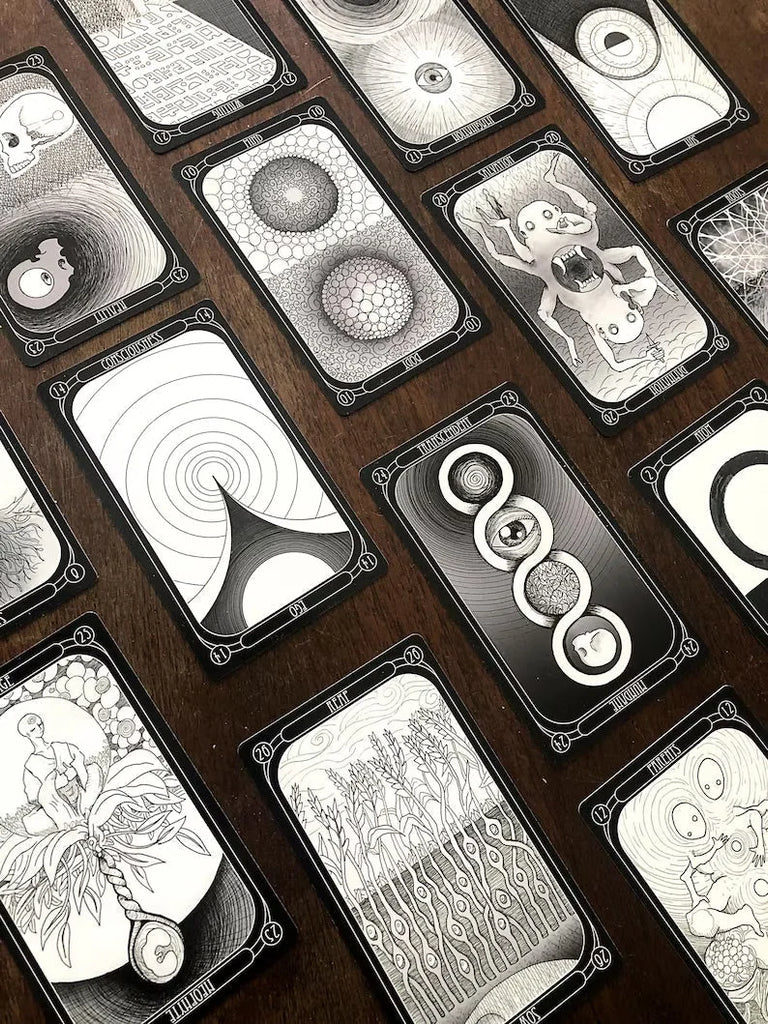 Collection of tarot cards fanned diagonally across dark wood, featuring black-and-white intricate illustrations of eyes, celestial symbols, organic patterns, and mystical imagery with ornate corner de