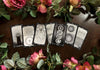 Seven tarot cards with intricate black-and-white geometric designs spread across dark wood, surrounded by fresh pink and coral roses with green foliage.