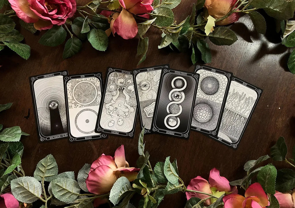 Seven tarot cards with intricate black-and-white geometric designs spread across dark wood, surrounded by fresh pink and coral roses with green foliage.