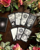 Seven tarot cards with intricate black and white eye and circular motifs arranged on dark wood, surrounded by pink roses and green foliage, with a dark card case visible at left.
