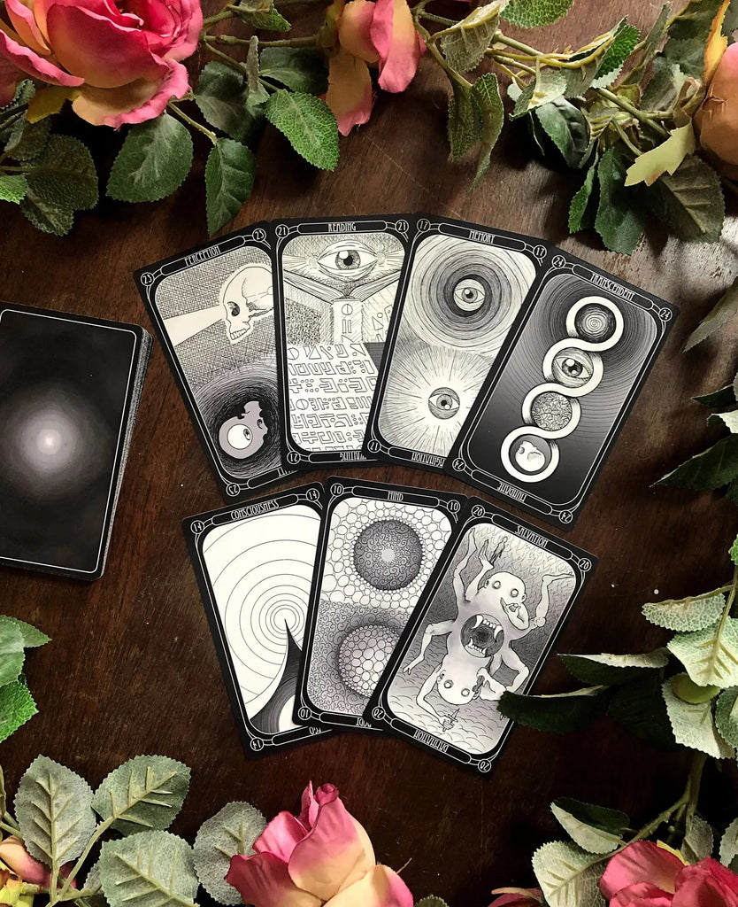 Seven tarot cards with intricate black and white eye and circular motifs arranged on dark wood, surrounded by pink roses and green foliage, with a dark card case visible at left.